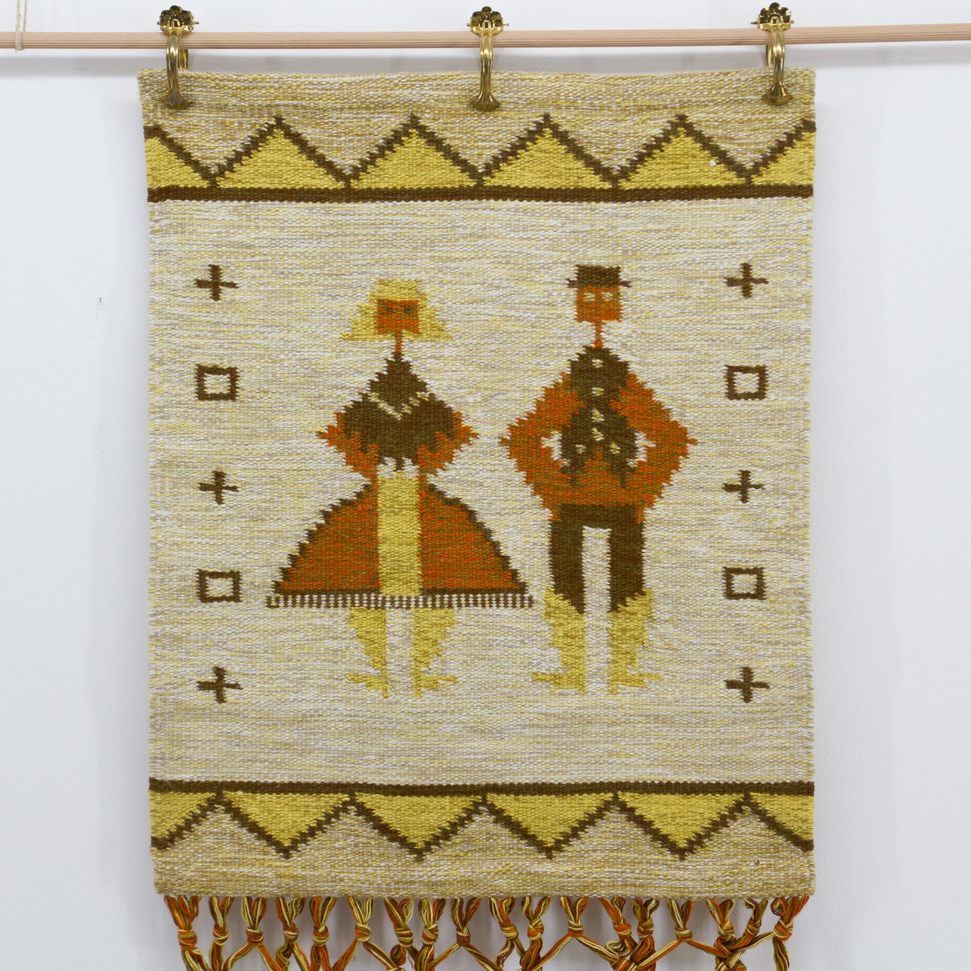 Straight-on, well-lit view focusing entirely on the beautifully woven flatweave section of the vintage folk tapestry, showing stylized geometric human figures in mustard yellow and terracotta, styled gracefully using brass hanging clips.