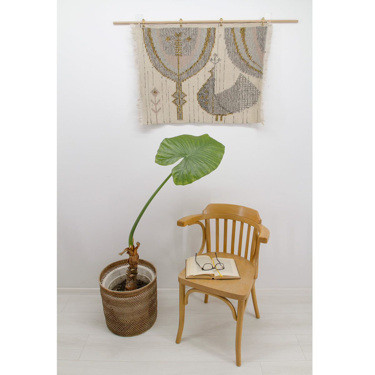 The vintage wall hanging styled vertically on the wall in a bright mid-century modern setting. It hangs on a wooden dowel above a bentwood chair and a large potted Alocasia plant, demonstrating its scale and how the soft grey and pink tones complement natural wood furniture and retro home decor.
