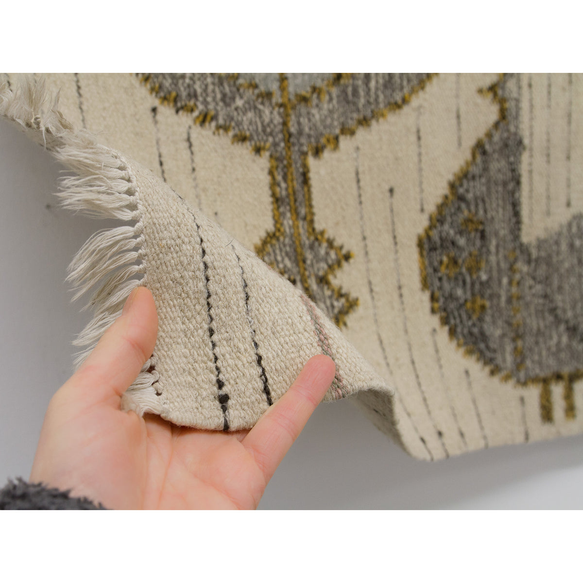 A hand holding the corner of the vintage tapestry to show the short, neat fringe finish. The softness of the cream wool is apparent, and the image confirms the hems are intact without unraveling.