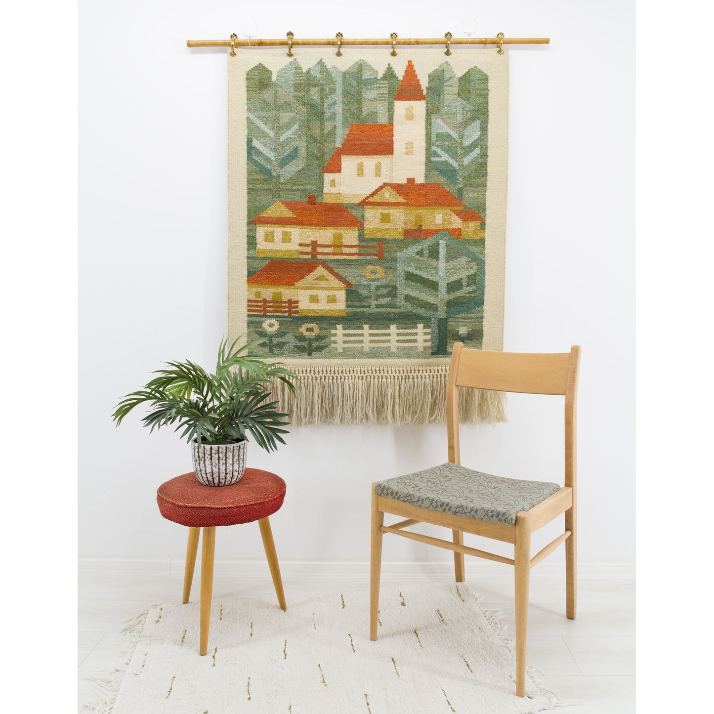 A large 1970s geometric village tapestry hanging on a bright wall behind a mid-century chair, featuring charming retro houses in forest green, red, and yellow.