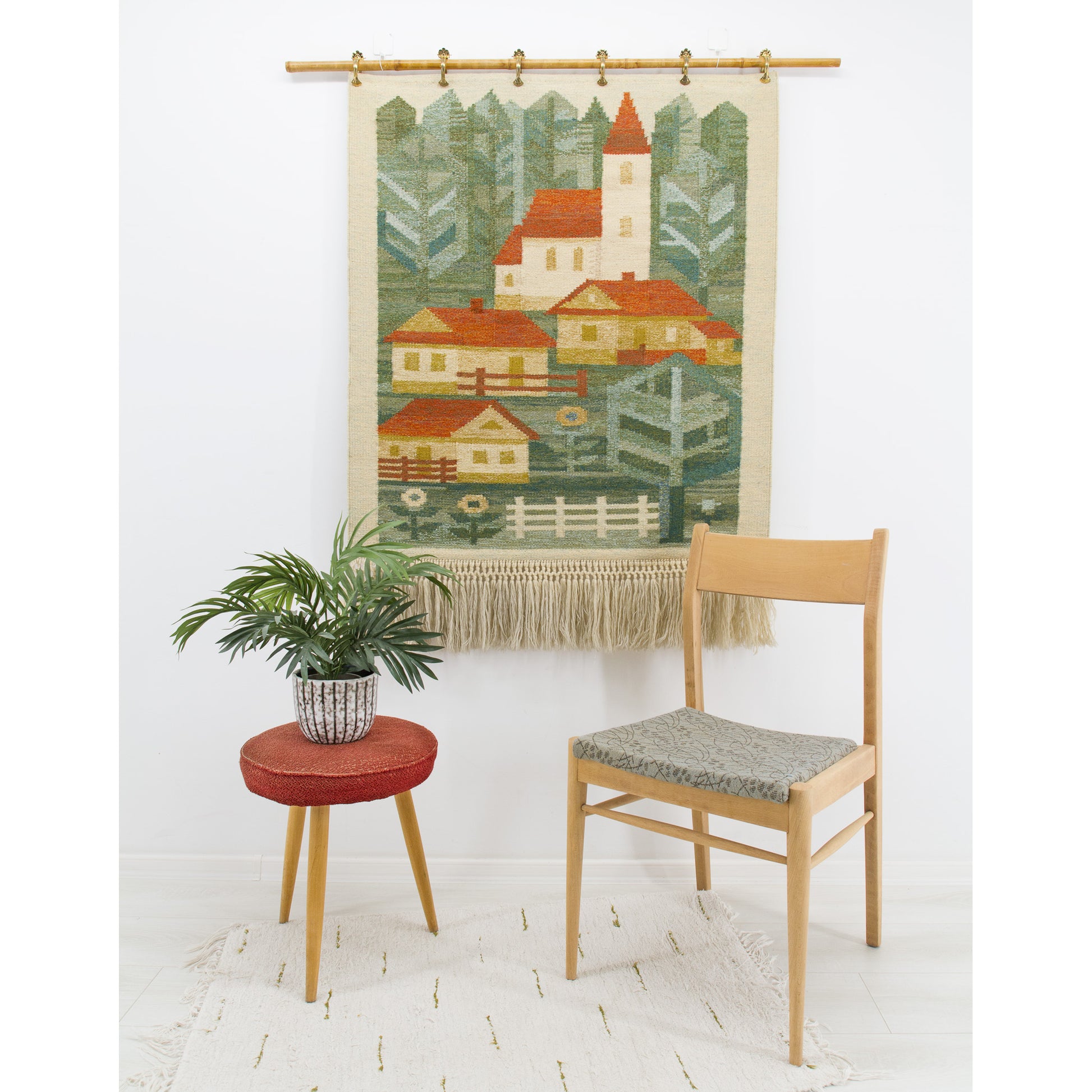 A large 1970s geometric village tapestry hanging on a bright wall behind a mid-century chair, featuring charming retro houses in forest green, red, and yellow.
