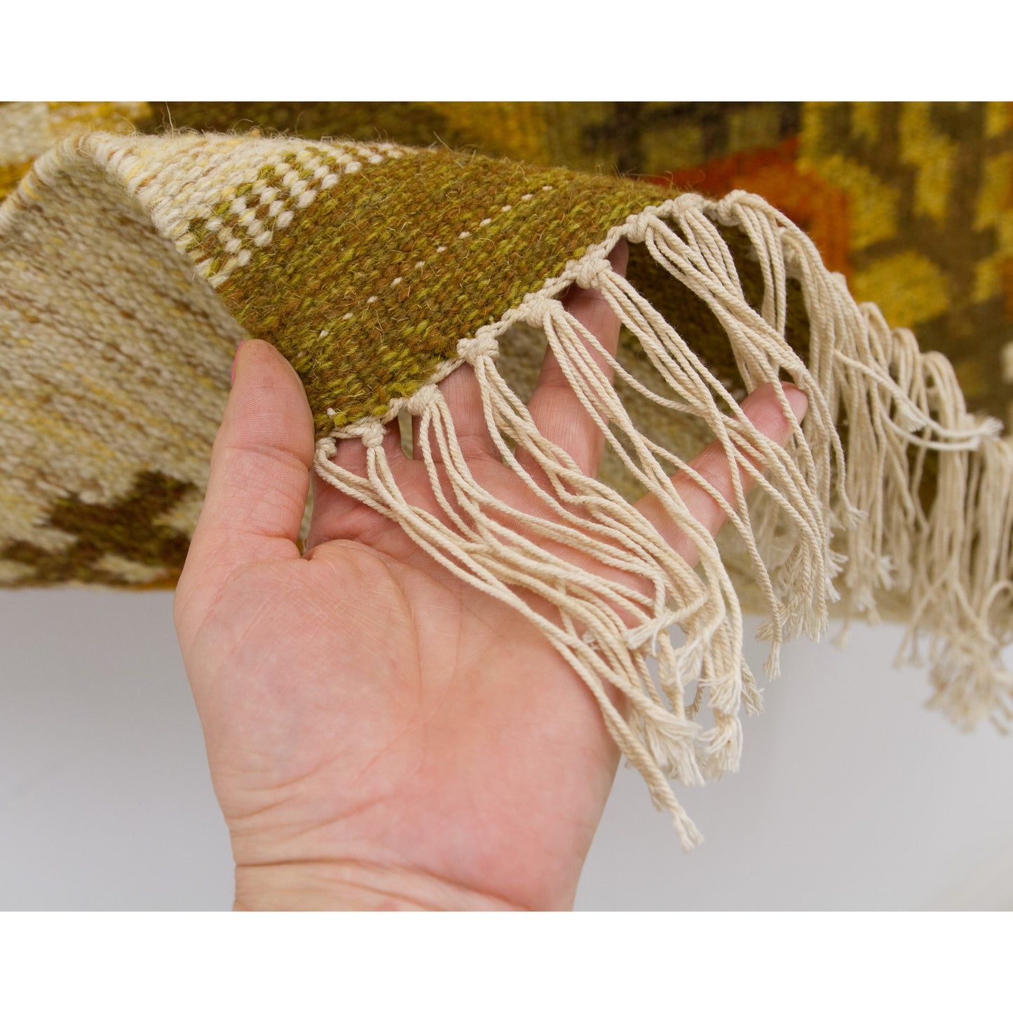 Hand lifting and showing the flawlessly finished row of neatly knotted four-inch soft yarn fringes dangling securely from the mid-century rug border.